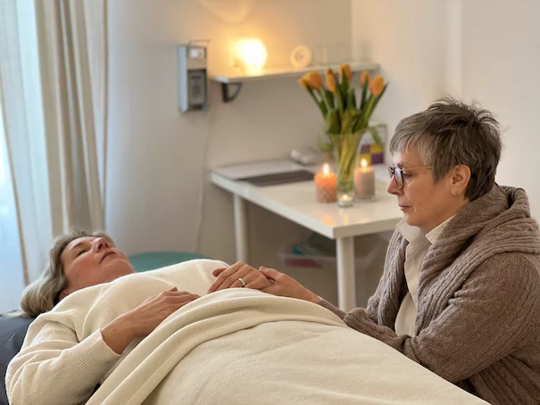Jin Shin Jyutsu Behandlung – Sanftes Strömen auf der Liege Jin Shin Jyutsu Behandlung in München: Julietta Jebens-Theimer hält sanft die Hände einer entspannt liegenden Klientin auf der Behandlungsliege